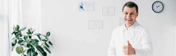 Панорамный снимок симпатичного улыбающегося хиропрактика, показывающего большой палец в больнице
