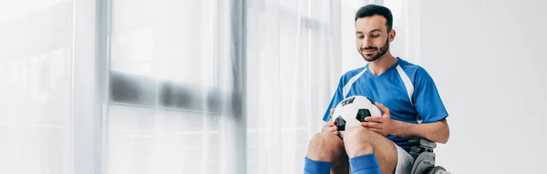 panoramic shot of man in football uniform sitting in Wheelchair and holding soccer ball