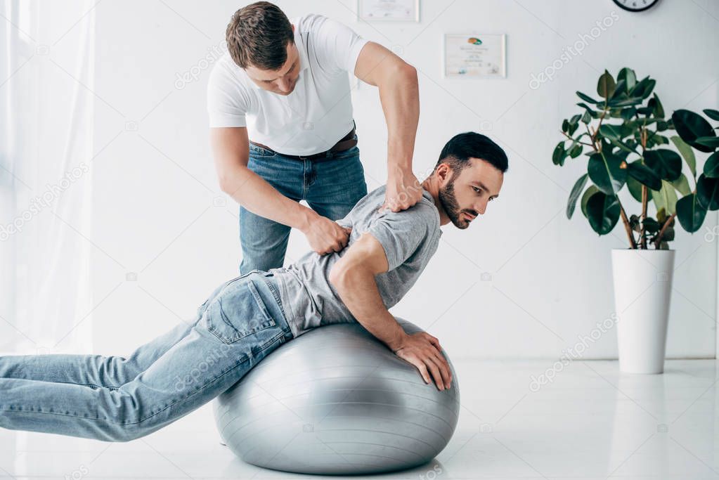 chiropractor massaging back of man lying on fitness ball in hospital
