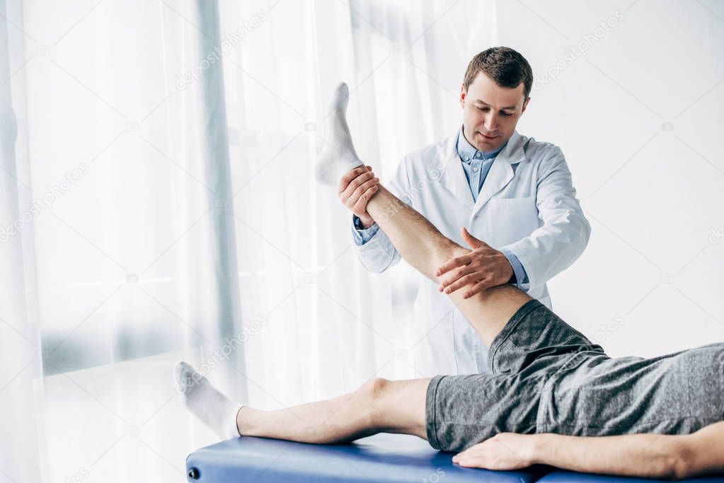 handsome Physiotherapist stretching leg of patient on massage table in hospital