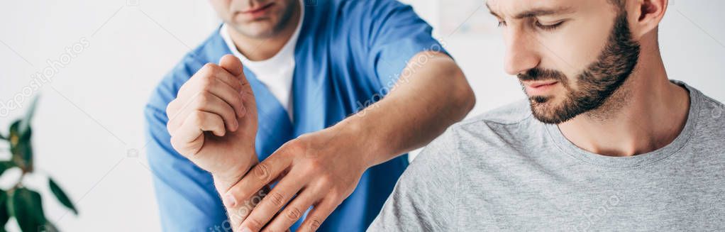 panoramic shot of chiropractor massaging arm of bearded patient in hospital