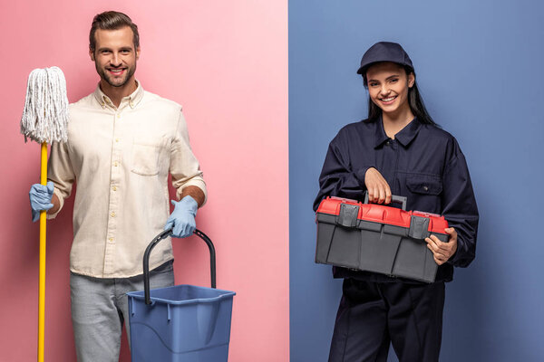 man in rubber gloves with mop and woman in construction worker uniform with tool box looking at camera on blue and pink