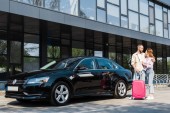 happy man hugging cheerful woman while standing near black car with pink luggage 