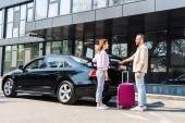 cheerful man looking at happy woman while standing near car and pink luggage 