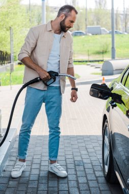 handsome man holding fuel nozzle near black car at gas station  clipart