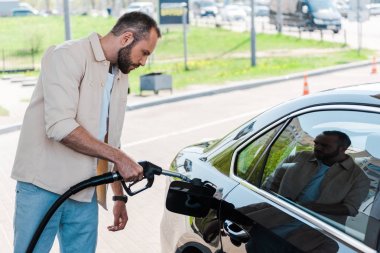handsome man holding fuel pump and refueling black car at gas station  clipart