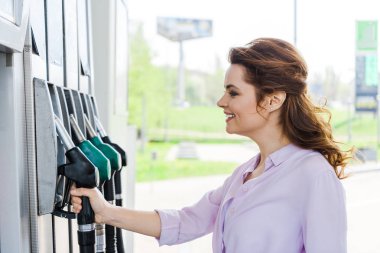 happy woman holding fuel pump while smiling at gas station  clipart