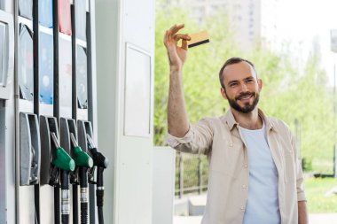happy bearded man holding credit card and smiling at gas station  clipart
