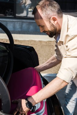 happy and bearded man smiling while putting pink luggage in car trunk  clipart