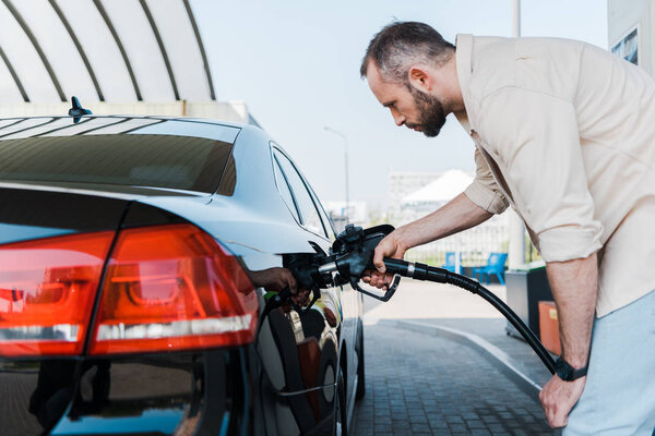 bearded man holding fuel pump and refueling black car at gas station 