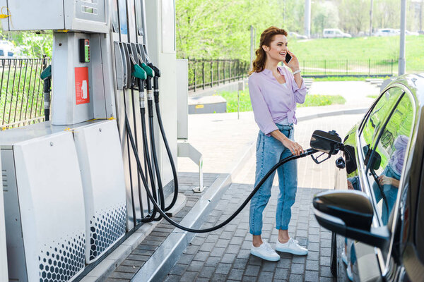 cheerful woman holding fuel pump while refueling car with benzine and talking on smartphone 