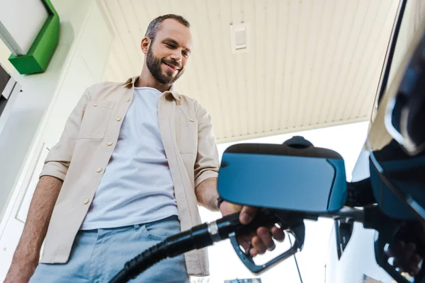 ガソリンスタンドでベンジンで自動車に燃料を補給しながら燃料ポンプを持つ幸せな男の低角度図 — ストック写真