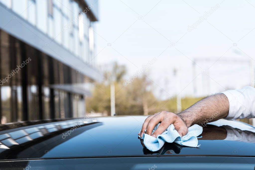cropped view of man cleaning black car with white cloth near building 