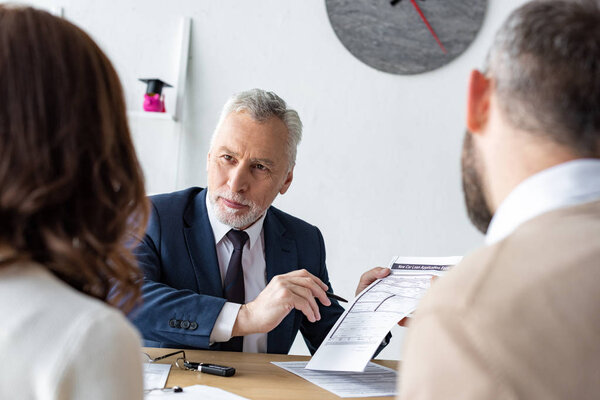 selective focus of bearded car dealer holding contract and pen near customers 