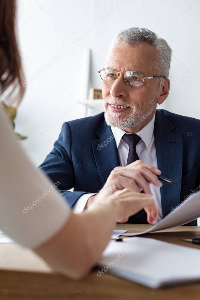 low angle view of happy car dealer in glasses holding pen and looking at woman in office 