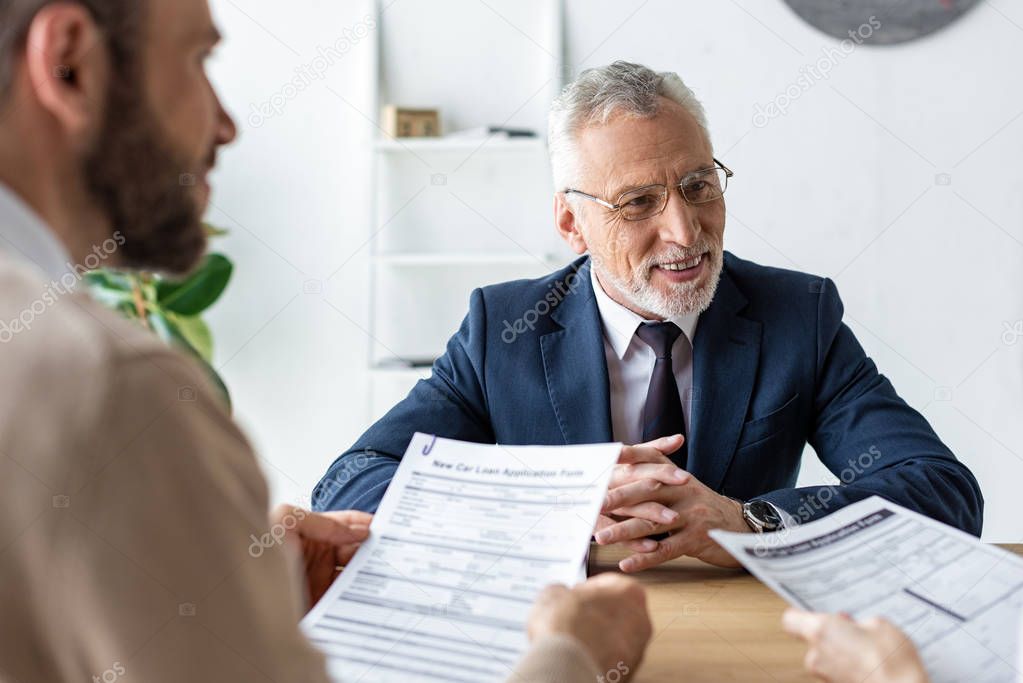 happy car dealer in glasses sitting with clenched hands near customers with contracts 