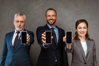 selective focus of cheerful businessmen and businesswoman holding smartphones with blank screens on grey  clipart