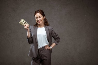 mutlu iş kadını kalça eliyle ayakta ve gri dolar banknotlar tutarak 