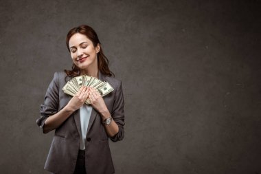happy businesswoman standing with closed eyes and holding dollar banknotes on grey  clipart
