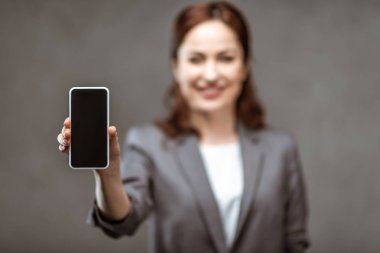 gri üzerinde boş ekran ile akıllı telefon tutan neşeli iş kadını seçici odak 