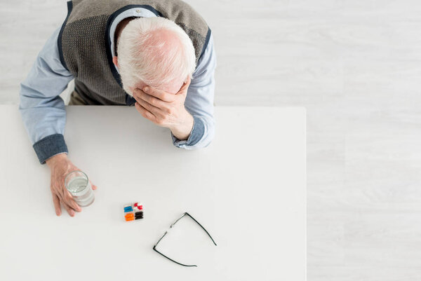 Overhead view of upset elderly man sitting behind table with pills and water in glass