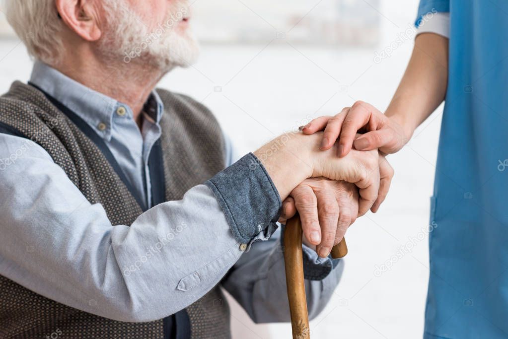 Cropped view of nurse holding hands with bearded senior man 