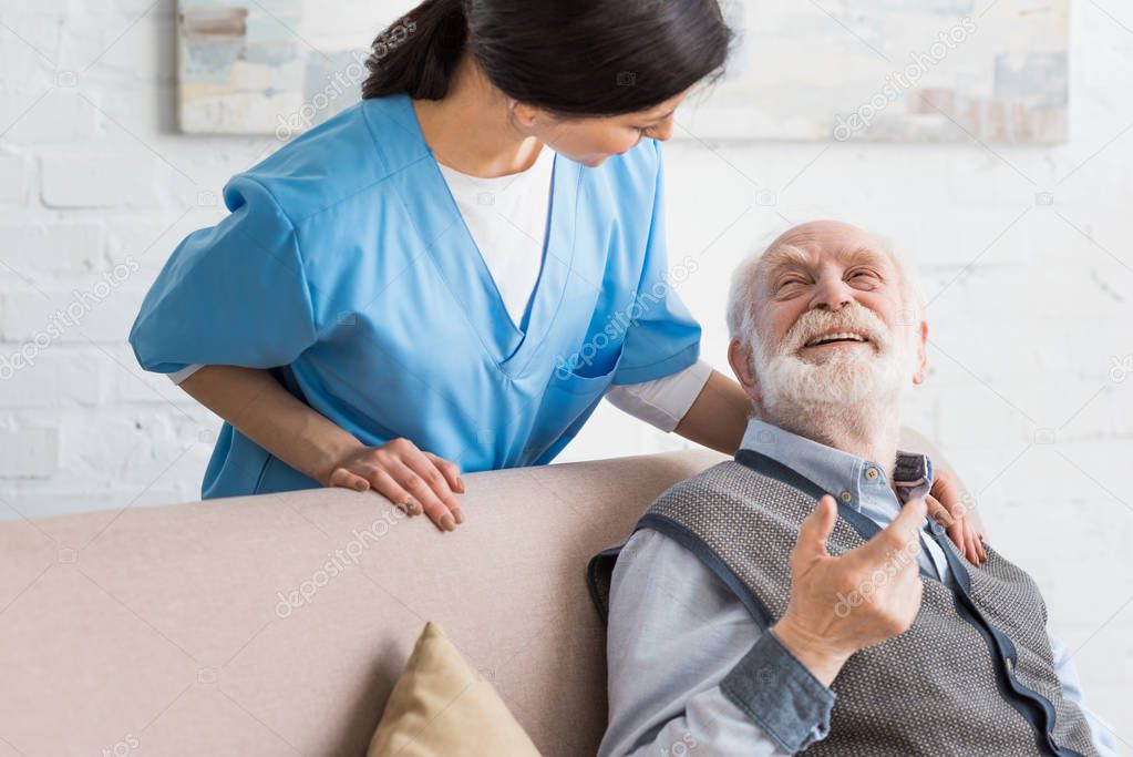 Happy senior man looking at nurse, sitting on couch