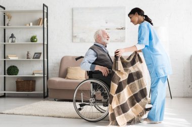 Side view of caring nurse, covering blanket disabled and grey haired man in wheelchair  clipart