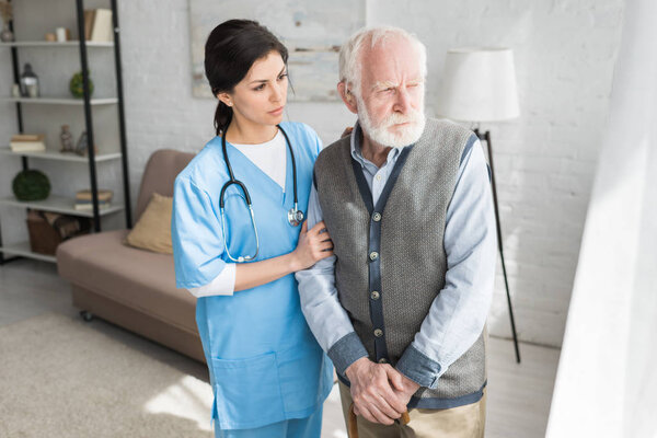 Doctor supporting of senior man, standing in bright light room, looking aside