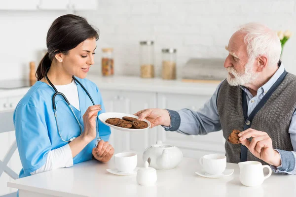 Homem Cabelos Grisalhos Oferecendo Biscoitos Para Feliz Enfermeira Sorrindo — Fotografia de Stock