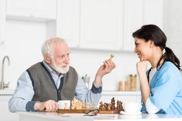 Krankenschwester Und Grauhaariger Mann Sitzt Auf Heller Küche Und Spielt — Stockfoto