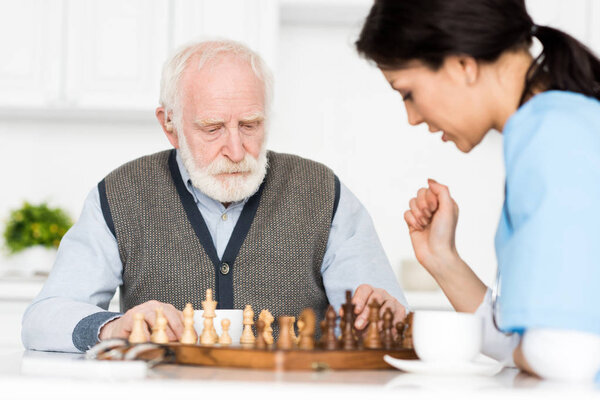 Selective focus on retired man sitting on kitchen, and playing in chess with nurse