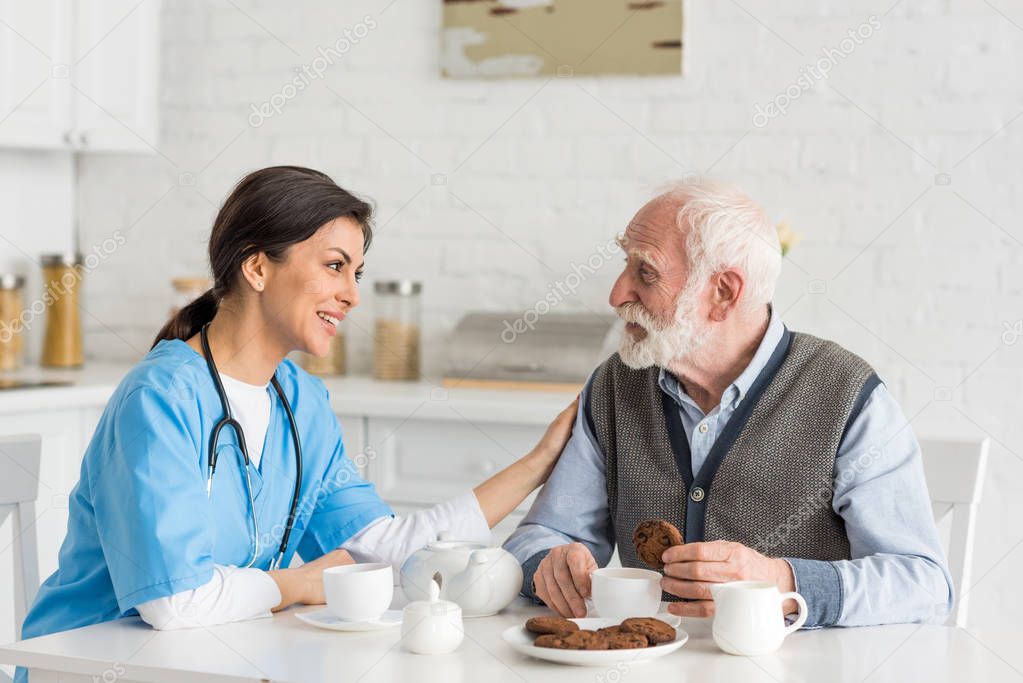 Nurse putting hands on grey haired man, sitting on kitchen