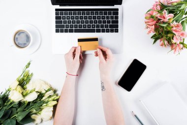 dizüstü bilgisayarın yanında kredi kartı tutan kadının kırpılmış görünümü, boş ekranlı akıllı telefon, beyaz bir fincan kahve ve buketler