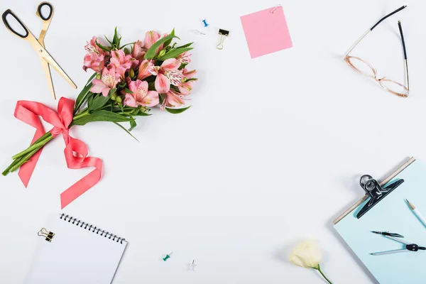 top view of flowers with ribbon, clipboard, scissors, notebook, pencil and glasses on white