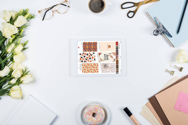 KYIV, UKRAINE - MAY 11, 2019: top view of cup of coffee, doughnut, flowers, glasses, book, papers, clipboard and digital tablet with pinterest app on screen on white