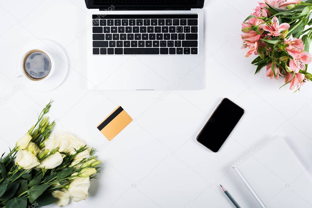 top view of laptop, smartphone with blank screen, credit card, book, pencil, cup of coffee and flowers on white