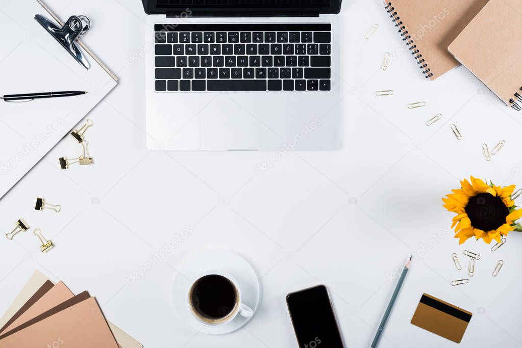 top view of laptop, smartphone with blank screen, sunflower, clipboard, cup of coffee, craft paper, credit card, binder clips and paper clips on white