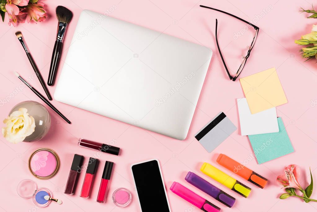 top view of laptop, smartphone with blank screen, credit card, glasses, flowers and decorative cosmetics on pink