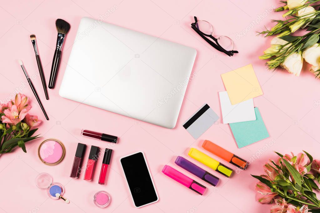 top view of laptop, smartphone with blank screen, credit card, flowers, glasses, highlighters and decorative cosmetics on pink