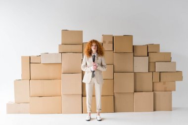 attractive curly redhead girl looking at camera while standing near boxes on white  clipart