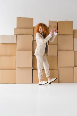 attractive curly redhead girl standing near boxes on white  clipart