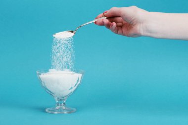 partial view of woman adding granulated sugar into glass bowl on blue background clipart