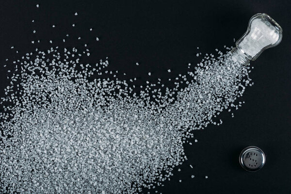 top view of salt crystals sprinkled near salt shaker on black background