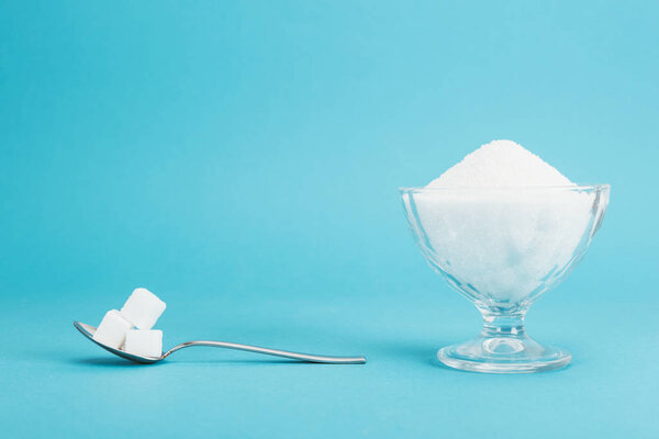 glass bowl with granulated sugar and teaspoon with sugar cubes on blue background with copy space