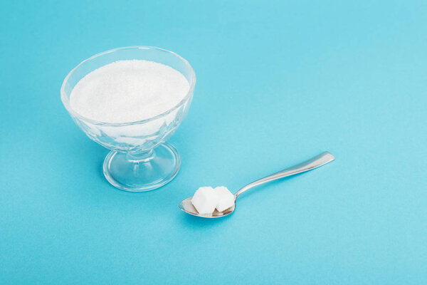 glass bowl with granulated sugar and teaspoon with sugar cubes on blue background 