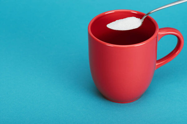 teaspoonful of white granulated sugar near red cup on blue background