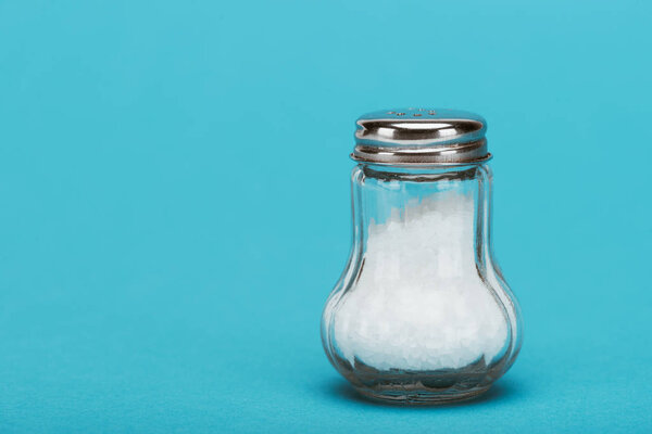 salt crystals in glass salt shaker with metallic cap on blue background with copy space