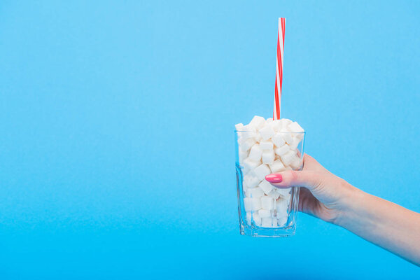 partial view of woman holding glass with straw and white sugar cubes isolated on blue