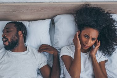 top view of african american woman suffering from headache while lying in bed near snoring husband clipart